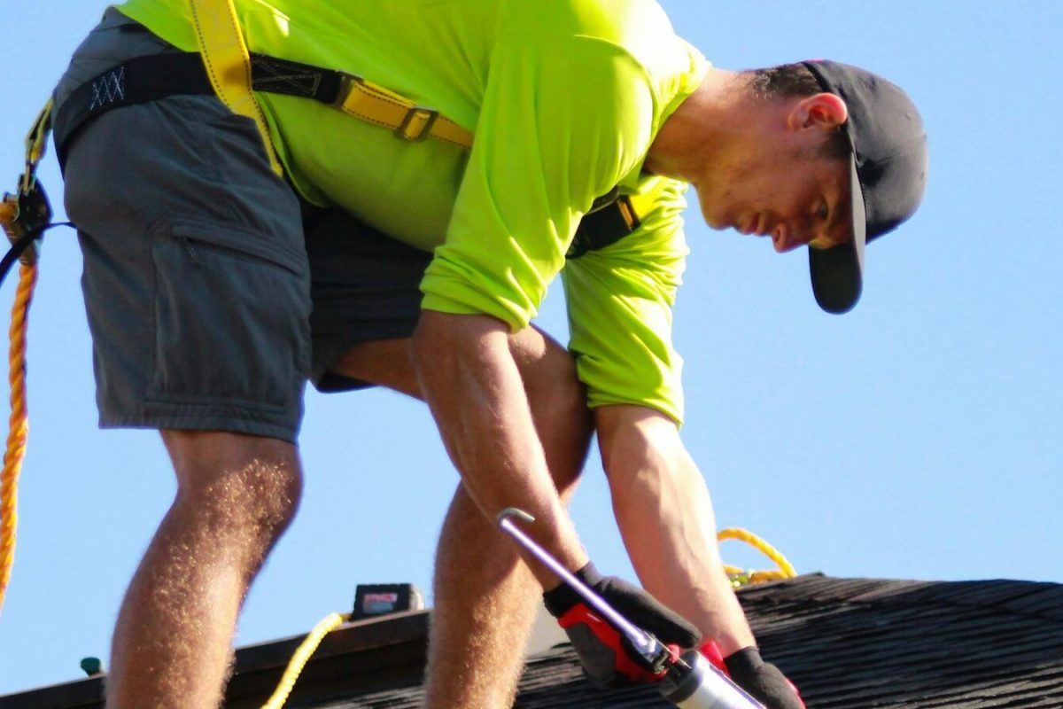 Roofer using a nail gun on an asphalt shingle roof against a blue sky